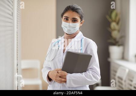 Portrait of female doctor in protective mask and in white coat holding medical cards in her hands looking at camera at hospital Stock Photo