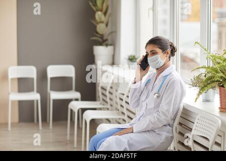 Female doctor in white coat and in protective mask sitting on chair and taking on mobile phone at hospital Stock Photo