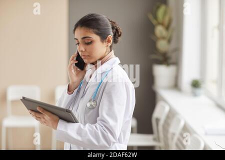 Female doctor in white coat has a conversation on mobile phone and looking at digital tablet in her hands standing at hospital Stock Photo