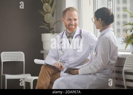 Smiling bearded doctor in white coat talking to young nurse while they sitting on chairs at the corridor Stock Photo