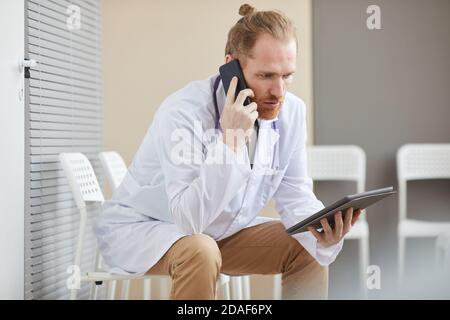 Male doctor has a conversation on mobile phone while using digital tablet sitting on chair at hospital Stock Photo