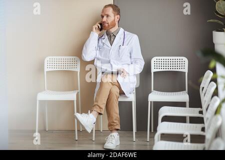 Male doctor in white coat sitting on chair talking on mobile phone and using tablet pc during his break at hospital Stock Photo