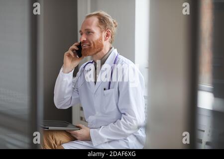 Confident doctor in white coat has a conversation on mobile phone while sitting at the corridor Stock Photo