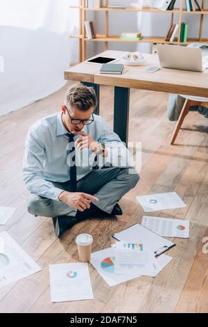 Pensive businessman looking at documents near coffee to go on floor in office Stock Photo