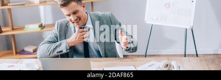 Smiling businessman having video chat on laptop near papers, cup and headphones on table, banner Stock Photo