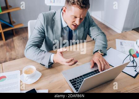 High angle view of angry businessman using laptop near cup of coffee and papers with charts on table Stock Photo