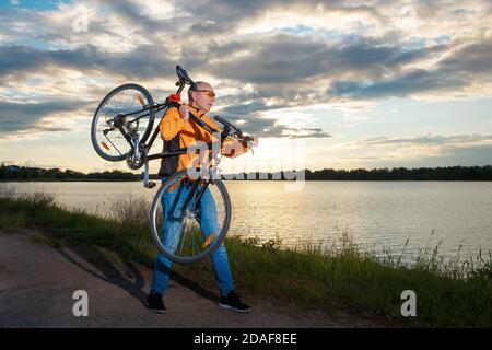 Tourist biker carries a bicycle on his shoulder against the backdrop of the setting sun on the lake. Stock Photo