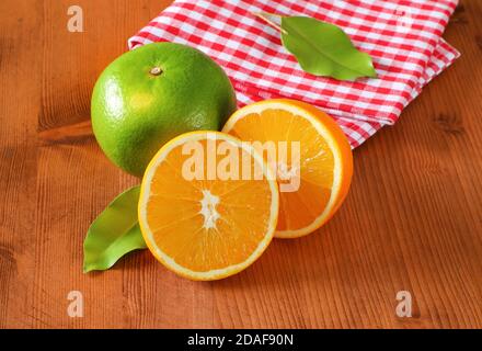 Green grapefruit (sweetie, pomelit, oroblanco), two orange halves and checked red and white tea towel on wooden table Stock Photo