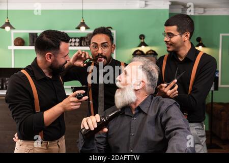Multiethnic barbers joke with a customer with a beer in hand. Alternative business.have barbering tools in their hands, such as scissors, shaving brus Stock Photo