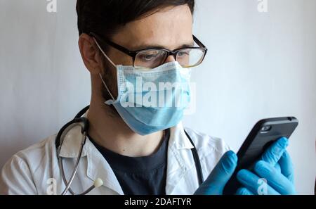 A young handsome doctor wearing glasses and a medical face mask provides a patient with a video chat consultation over the phone. Stock Photo