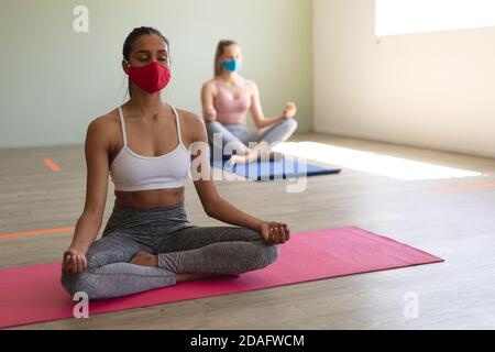 Two fit caucasian woman wearing face masks practicing yoga while sitting on yoga mats in the gym Stock Photo