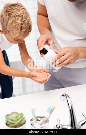 Father squeezing liquid soap on hand of son leaning forward, while standing on chair in bathroom on blurred foreground Stock Photo