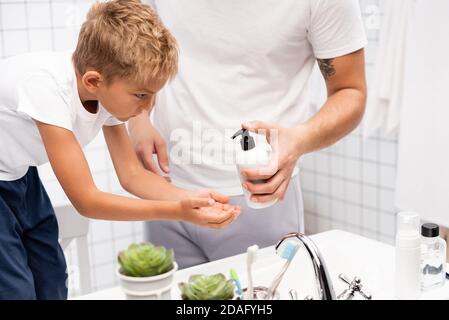 Man squeezing liquid soap on hand of boy leaning forward, while standing on chair in bathroom Stock Photo