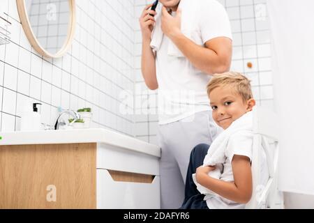 Positive boy looking at camera while sitting on chair near man shaving with electric razor in bathroom Stock Photo