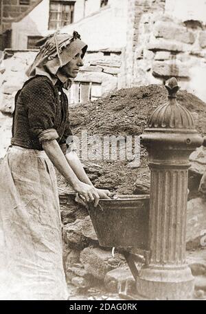 A Victorian photograph of a Woman collecting water at the Cragg Pump, Whitby, North Yorkshire in the late 1800s. Steps led down from the pump onto Pier road where fishwives would 'gip' or gut herrings as they arrived on the quay directly from the fishing boats. The pump was then the only source of fresh water in the area. The headress she is wearing is known as a 'Steerths' Bonnet, Taking its name from the village of Staithes where they were manufactured, though they were widely worn amongst the fishing communities elsewhere. Stock Photo