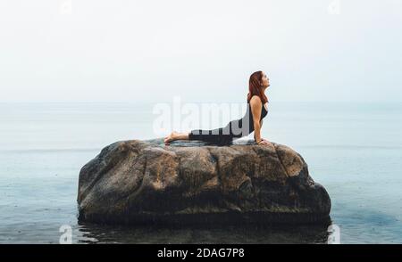 Caucasian young woman practicing yoga alone, cobra pose, on a rock, in the Baltic Sea, in the rain. Red hair girl meditating in seascape, on rainy day Stock Photo