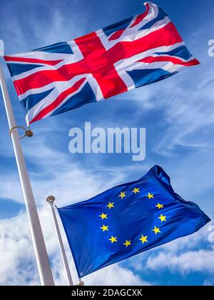 BREXIT FLAGS EU CONCEPT UK Union Jack Flag flying high above EU European Flag in a stiff breeze on a sunlit day with blue sky & flags surrounded by swirling clouds Stock Photo