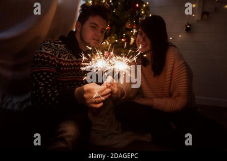 Stylish happy couple holding burning sparklers on background of  christmas tree with lights in dark festive room. Young family celebrating new year wi Stock Photo