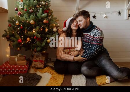 Stylish happy couple holding christmas gift under christmas tree with  lights in festive decorated room. Young family exchanging presents and hugging, Stock Photo