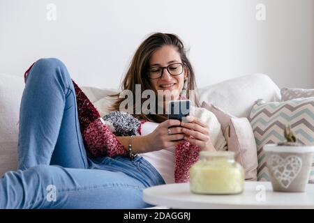 Smiling young woman at home relaxing in the sofa with a cell phone in her hands. Stock Photo