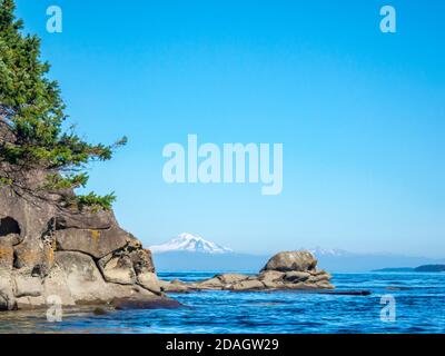 A reef extends out from an eroded island shoreline in the Strait of Georgia, framing a view of Mt. Baker, whose snowy peak rises in the distance. Stock Photo