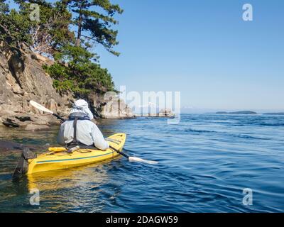A man, dressed for protection from the summer sun, paddles a yellow kayak in BC's southern Gulf Islands, with Mt. Baker's snowy peak in the distance.