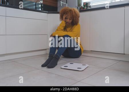 Devastated young african american black woman sitting on the flor with weight scale in front. Weight loss and mental health concept. High quality photo Stock Photo