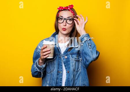 Young woman in denim jacket, headband and glasses holding paper cup in hands on yellow background