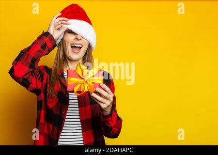 Young woman peeks out from under a santa hat and a red checkered shirt and holds a gift in her hands on a yellow background Stock Photo