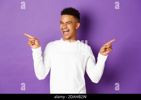 Image of cheerful african-american guy in white sweatshirt, showing two variants, pointing fingers sideways at copy space and smiling, demonstrate Stock Photo