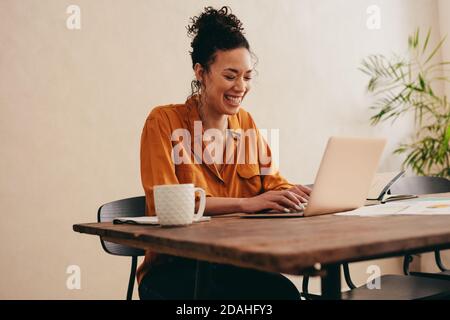 Smiling woman working on laptop at home. Female sitting at table with a coffee cup, using laptop. Stock Photo