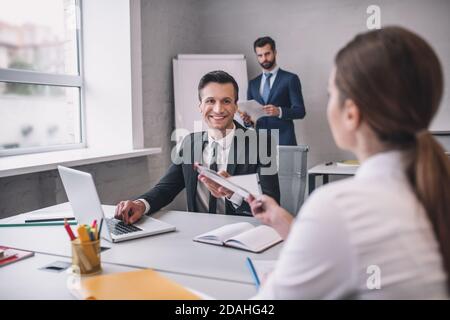 Smiling man with laptop taking documents from woman Stock Photo