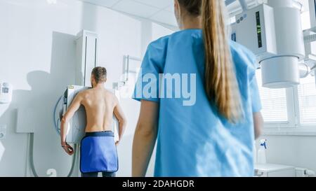 In the Hospital, Man Standing Face Against the Wall While Medical Technician Looking at Him Before X-Ray Scanning. Scanning for Fractures, Broken Stock Photo
