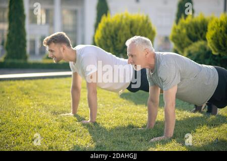 Young brown-haired male and mature grey-haired male doing push ups on grass Stock Photo