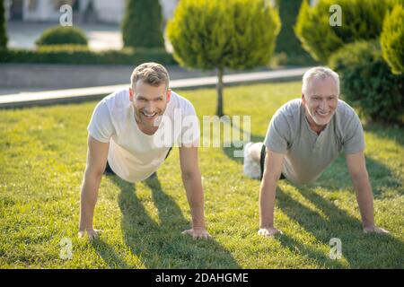 Young male and grey-haired male doing push ups on grass, smiling Stock Photo