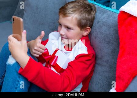 Handsome kid boy in Santa Claus hat looks at smartphone screen, shows thumb up.Talk with family,closeup image. Stock Photo