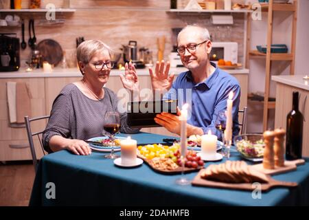 Mature man and woman waving at tablet pc during video conference in kitchen celebrating relationship. Couple sitting at the table, browsing, talking, using internet, celebrating their anniversary in dining room. Stock Photo