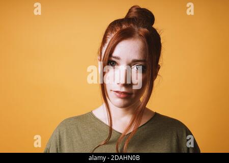 cool young woman with red hair messy bun hairstyle and loose strands at the front against yellow orange color background with opy space Stock Photo