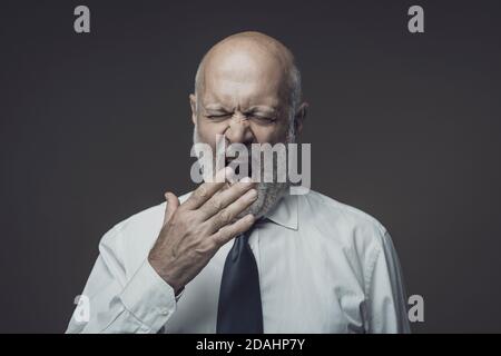 Lazy tired middle-aged businessman yawning and feeling sleepy, he is covering his mouth with his hand Stock Photo
