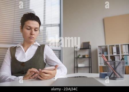 Serious young woman sitting at the table at office she typing a message using her mobile phone Stock Photo