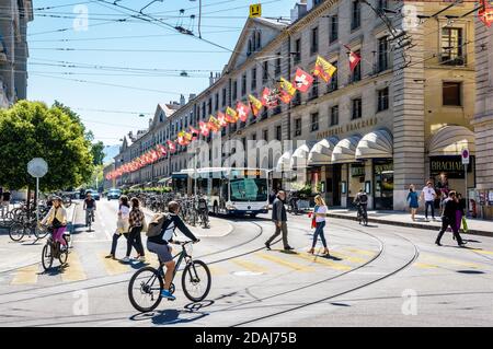 Pedestrians and cyclists are crossing the rue de la Corraterie in Geneva, with buildings decked with flags, as a bus is waiting at a red light. Stock Photo