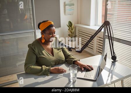 African radio host sitting at the table typing on laptop while speaking on microphone during her work on radio Stock Photo