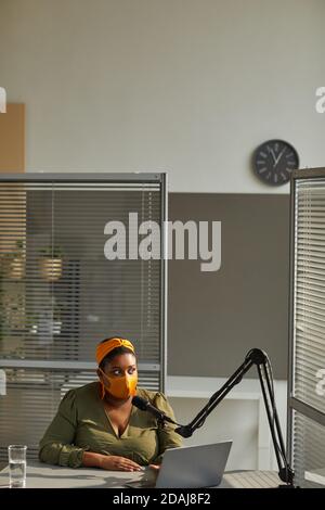 African radio dj in protective mask sitting at the table with laptop and speaking on microphone Stock Photo