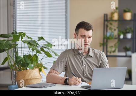 Young office worker sitting at his workplace looking at computer monitor and making notes in note pad Stock Photo