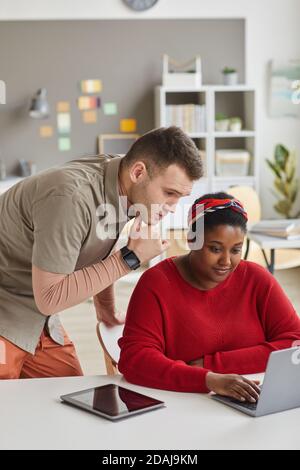 African woman sitting at the table and watching something on laptop with man talking to her they working at office Stock Photo