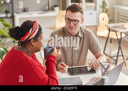 Two colleagues in casual clothing sitting at the table with tablet pc and coffee and talking to each other Stock Photo