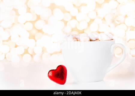 Gift card with chocolate red heart in red foil white cup of coffee and marshmallow on golden background of christmas lights bokeh. Mockup for decorative design. Christmas and New Year concept. Copy space. Stock Photo