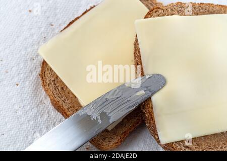 Overhead close view of a butter knife atop two slices of wheat bread with margarine and cheese on paper towels illuminated with natural light. Stock Photo