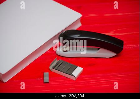 Black office stapler i Stationery for paper stitching. Stapler and staples with sheets of paper on a wooden red table. Stock Photo