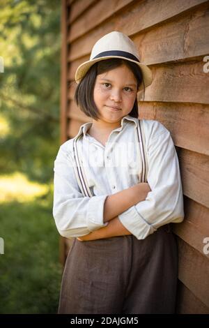 Country girl poses leaning against wooden plank wall of building. Teenage girl in white hat and braces looks at camera crossed arms. Holidays in Stock Photo